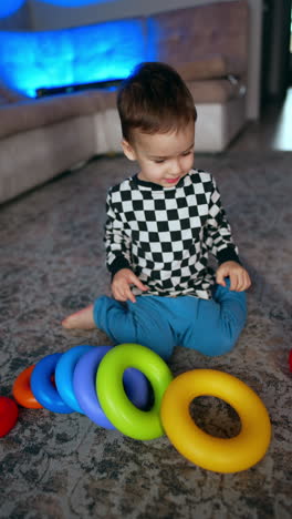 young baby boy holding a colorful pyramid while sitting on the floor. kid disassembles the toy and starts to build again. vertical video.