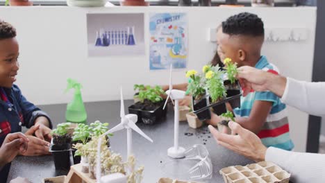 diverse male teacher and happy schoolchildren studying plants and wind turbines in school classroom