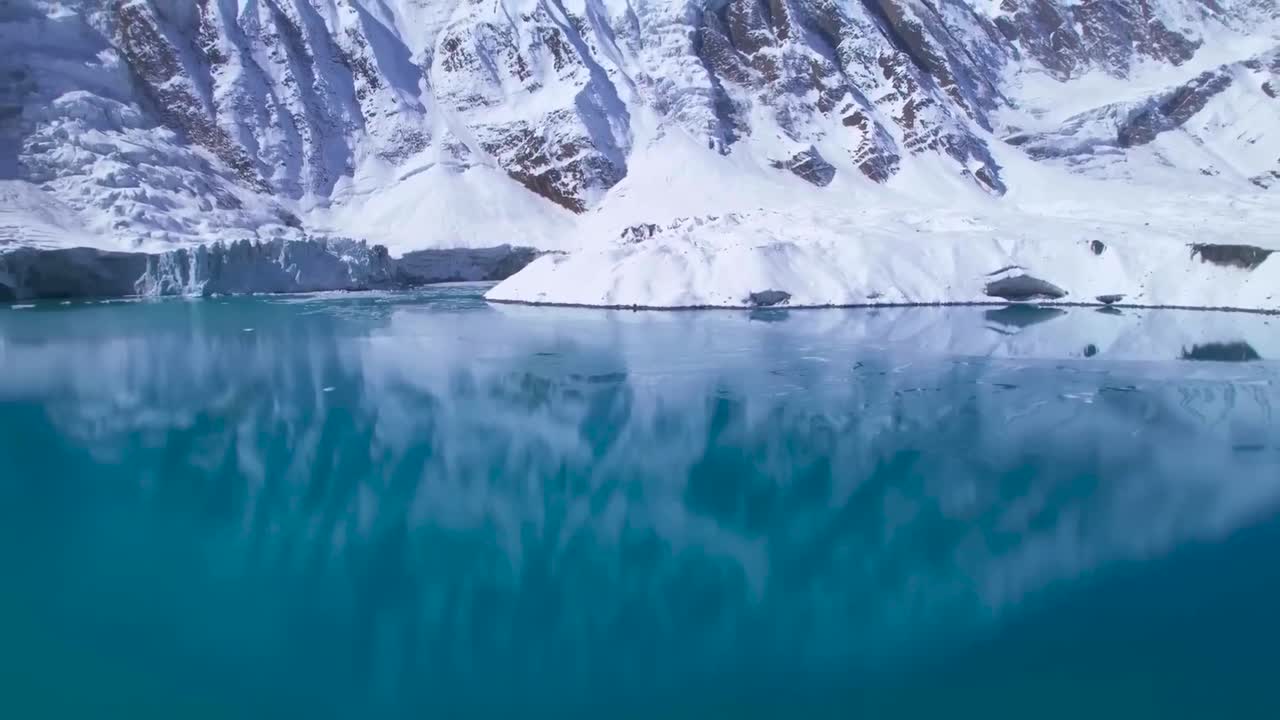 Aerial drone footage of blue icy and reflective Tilicho lake in Nepal during a sunny day while footage rises up slowly over the cold water that is reflecting the large mountains on the surface in back