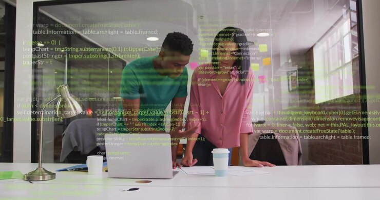 Code overlay appearing, two colleagues leaning, pointing, consulting laptop papers while reviewing