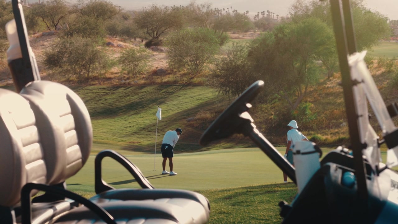 Un golfista esegue un putt su un green nel deserto inquadrato attraverso un golf cart mentre il sole tramonta, ripreso al rallentatore, adatto al lifestyle del golf e al marketing dei country club