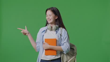 녹색 스크린 배경 스튜디오에 서 있는 동안 미소 짓고 으로 가리키는 뒷면과 몇 권의 책을 가진 아시아 소녀 학생