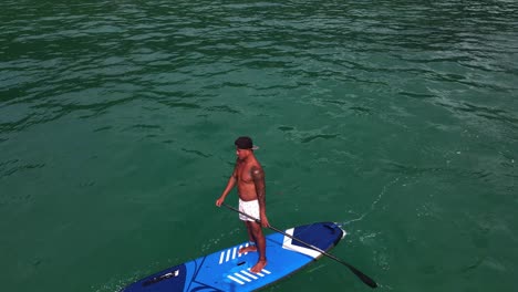 Aerial-drone-bird's-eye-view-of-a-Thai-man-exercising-on-a-sup-paddle-board-in-turquoise-tropical-clear-waters,-in-Thailand