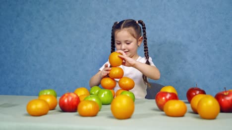 cute little girl with fresh fruit. funny girl building a tower of oranges on the table. child playing with organic fruit on blue background.