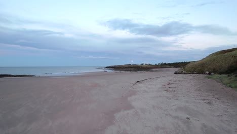fast moving drone footage flying close to the surface of a long sandy beach before rising up high above it towards the rising moon while the tide laps the shore