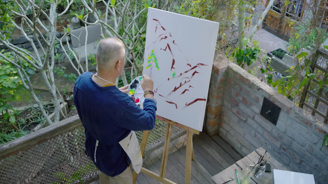 アジア系の高齢男性がパレットに筆を浸し、緑の木を描いている。デッキに置かれたキャンバスには、絵の具の隆起がアートを形作っている。