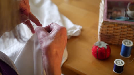 the hands of an aging elderly woman sewing a button on a white dress shirt by hand with a pincushion and spools of thread