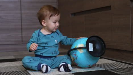 curious baby boy playing with a big globe. caucasian kid sitting on the floor rolls the sphere. mom comes up and puts the globe closer to her smiling son.