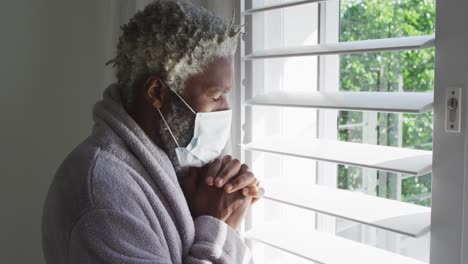 senior man wearing face mask looking out of window at home