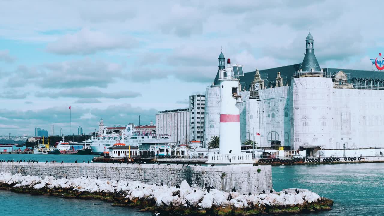 Flight around modern white and red lighthouse in Istanbul harbour with flying seagulls. Mediterranean sea calm waves many concrete buoys pier breakwater navigation shipping port City landscape skyline