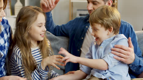 Cerca-De-Los-Niños-Pequeños-Y-Lindos-Divirtiéndose-En-El-Sofá-De-La-Sala-De-Estar,-Haciéndose-Cosquillas-Y-Riendo-Mientras-Sus-Padres-Los-Miran