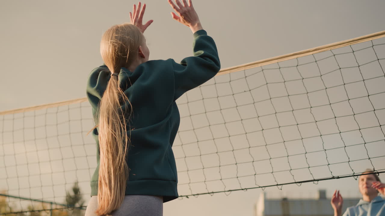 머리카락이 뒤로 인 녹색 후디를 입은 배구 선수의 뒷면, 상대가 잘못 치는 동안 공을 치려고 시도, 건물 배경과 은 하늘과 함께 야외 배구 경기