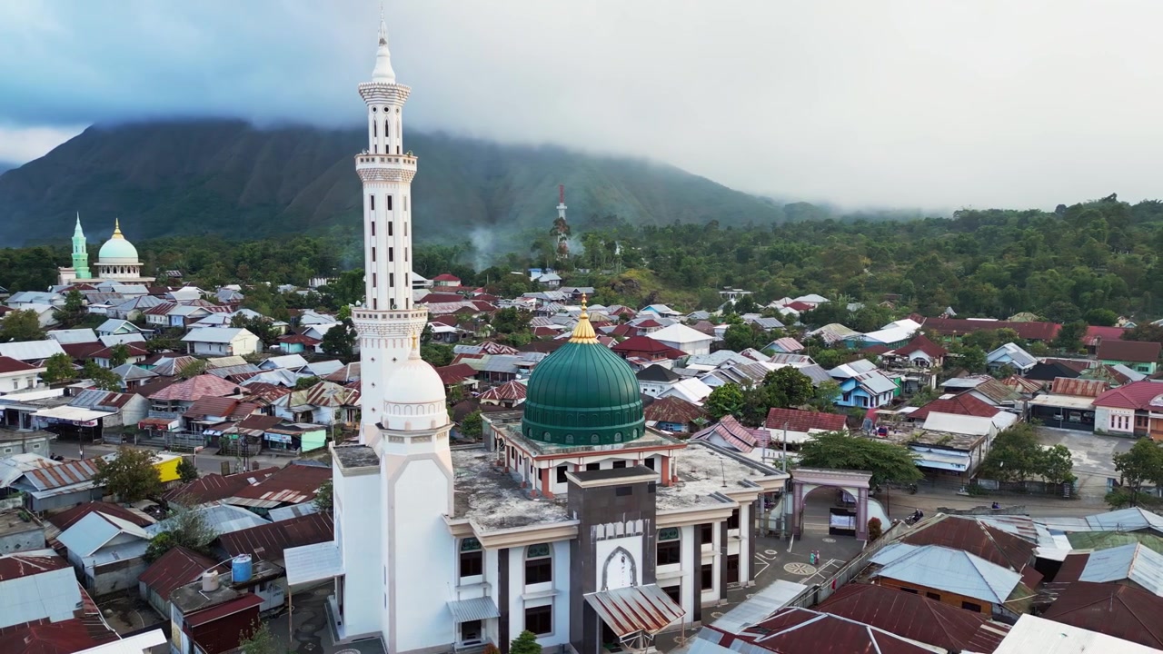Lingkungan pedesaan padat mengelilingi Masjid megah dengan rumah-rumah berkelompok di bawah pemandangan gunung dramatis dan lanskap lembah subur, rekaman drone udara.