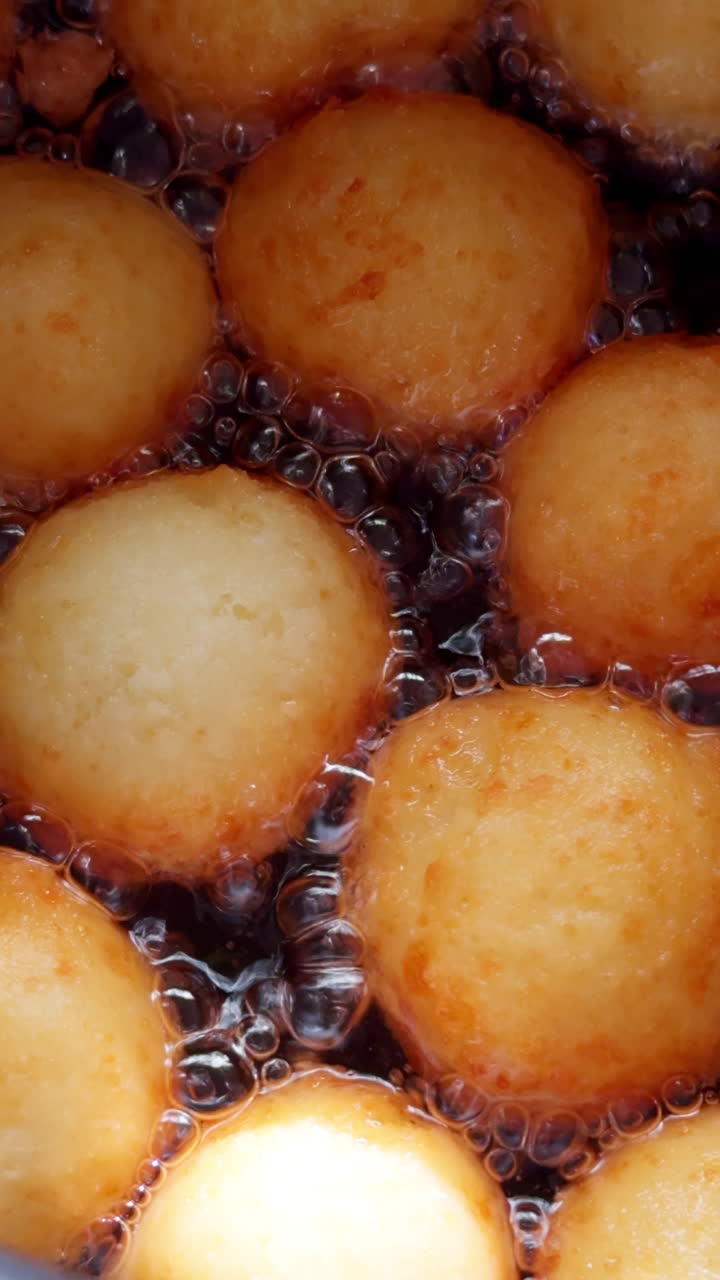 Delicious donuts with no central hole are turning gold in the boiling oil. Cooking sweet dough pastry in progress. Close up. Vertical video.