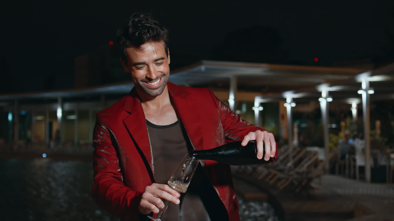 un tipo sonriente de fiesta nocturna junto a la piscina vertiendo champán en una copa de vidrio de cerca.