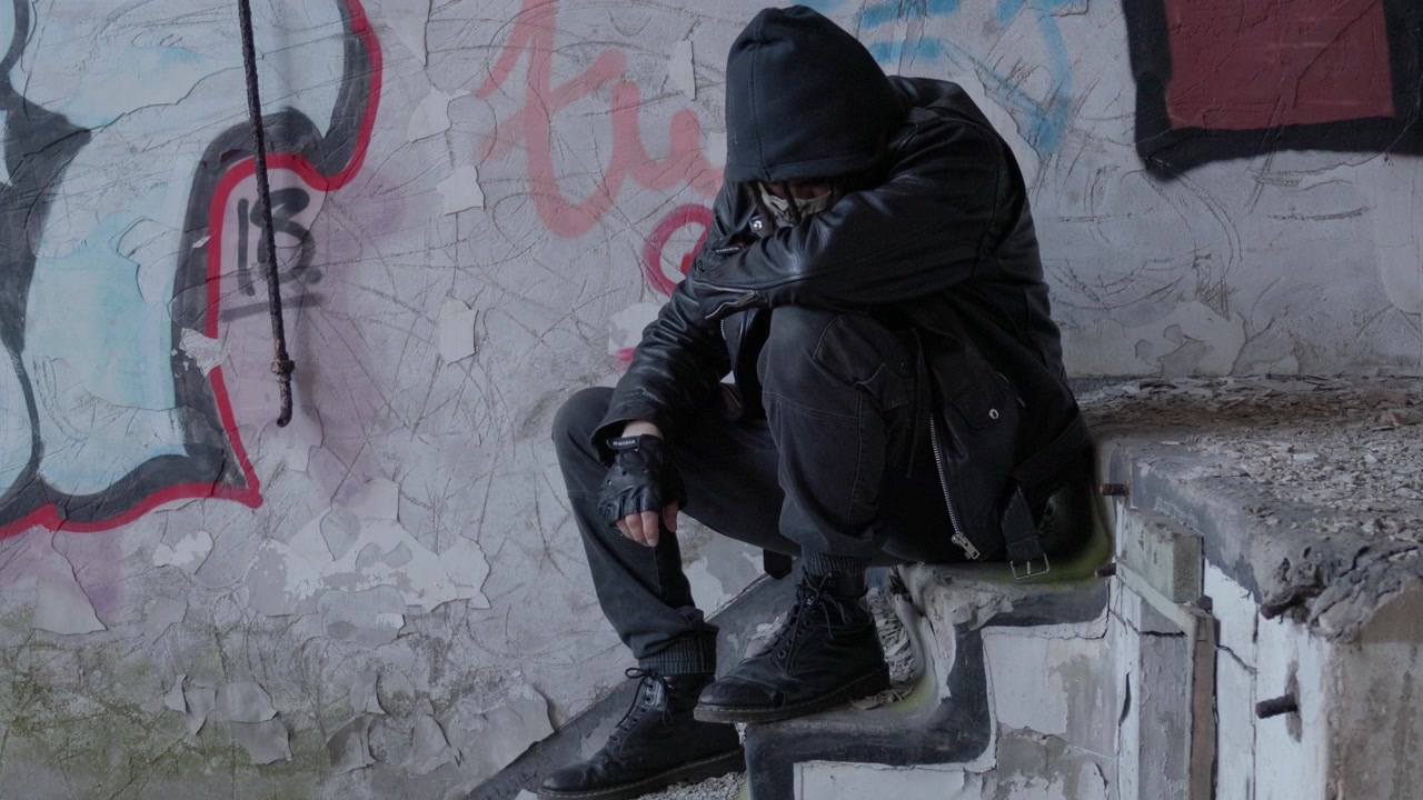 Una figura scura incappucciata siede da sola all'interno di una struttura abbandonata e coperta di graffiti, creando una scena suggestiva di isolamento, mistero e degrado urbano, ripresa statica con treppiede.