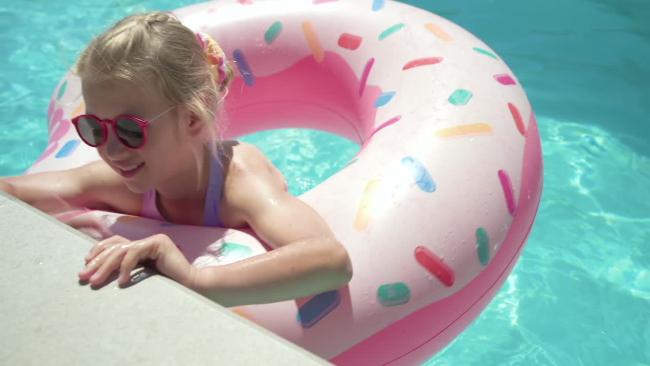 Female child in purple swimsuit smiling paddling and guiding donut float to reach yard pool coping