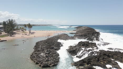 Drone-shot-of-a-bay-with-waves-crashing-over-top-revealing-beach-with-people-in-Puerto-Rico