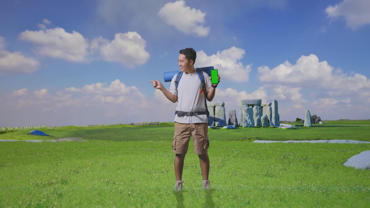 Full Body Of Asian Male Hiker With Mountaineering Backpack Showing Mock Up Green Screen Smartphone And Pointing To Side While Traveling In Stonehenge
