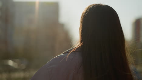 close-up of a girl with long brown hair flowing, wearing a peach jacket, walking through an urban area at sunset. the warm light highlights her hair as she turns her head slightly