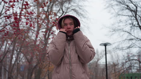 woman bundled in warm winter clothing adjusts her hoodie while walking through a snowy park, she holds her hands near her face to keep warm, surrounded by bare trees with red berries