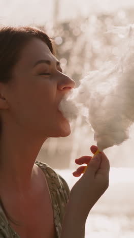 pretty lady eats sugar cotton wool against blurry fountain at sunset time closeup slow motion. street dessert in urban park. woman remembers carefree childhood