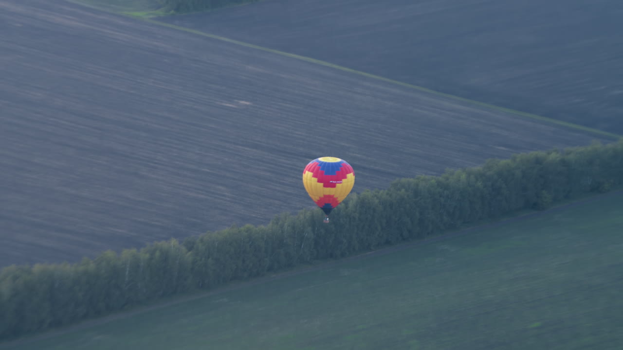 Distant view of vibrant hot air balloon descending over expansive tilled and grassy farmland, wreath of trees marking boundary as wicker basket sways beneath patterned canopy in tranquil