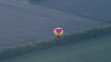 distant view of vibrant hot air balloon descending over expansive tilled and grassy farmland, wreath of trees marking boundary as wicker basket sways beneath patterned canopy in tranquil