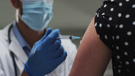 Caucasian-male-doctor-wearing-face-mask-giving-vaccination-to-a-female-patient