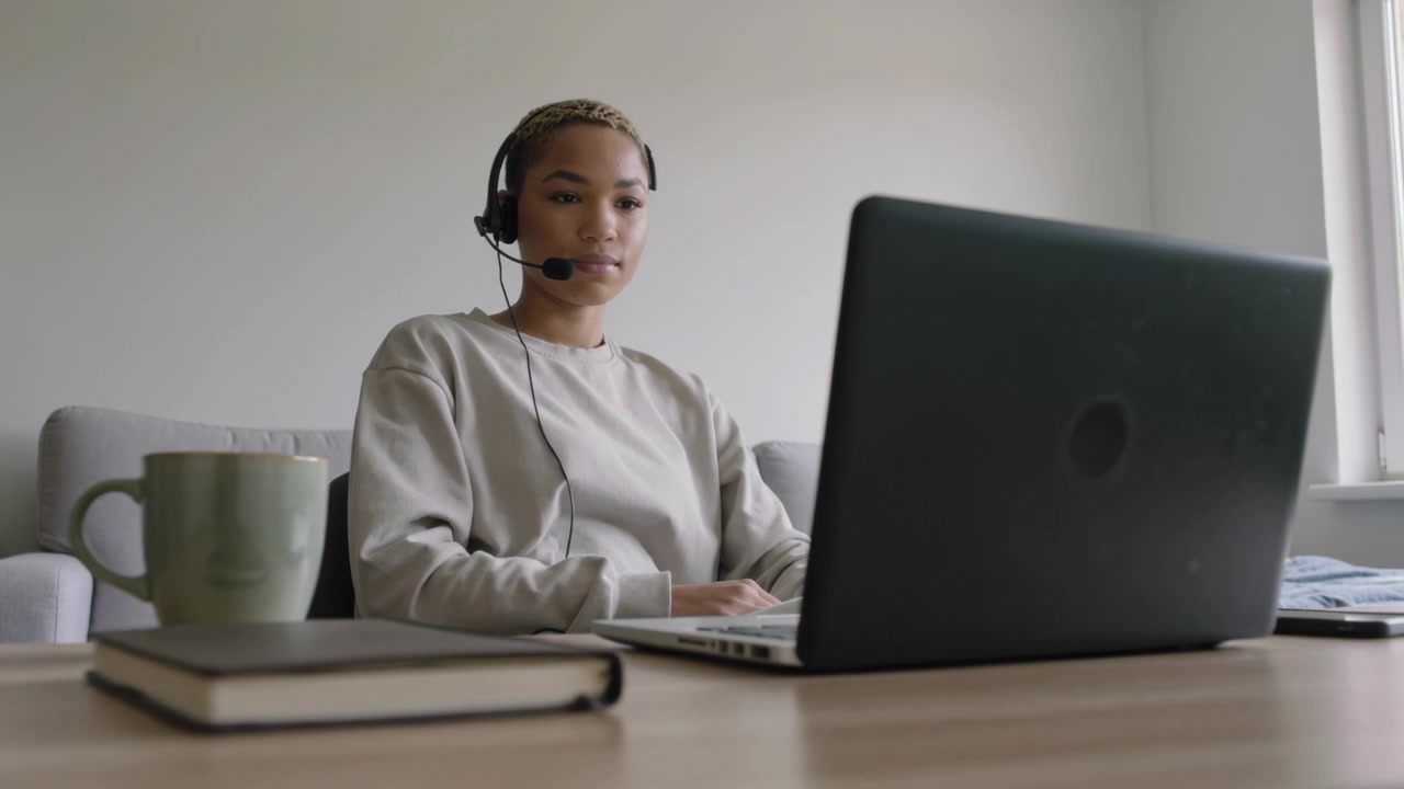 Mujer teletrabajando con sudadera gris, escribiendo en el portátil en casa, con auriculares, taza y cuaderno.