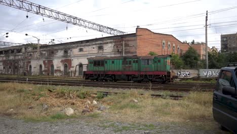 Green-old-locomotive-pass-in-front-of-old-crumbling-buildings-in-Tbilisi-Georgia,-graffiti-wall-in-the-background