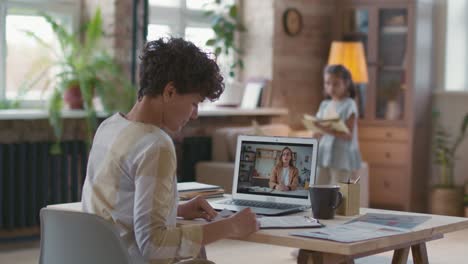 Frau-Mit-Kurzen-Locken-Sitzt-An-Einem-Schreibtisch-Vor-Dem-Computer-Und-Stellt-Ein-Projekt-Vor,-Im-Hintergrund-Liest-Seine-Tochter-Ein-Buch