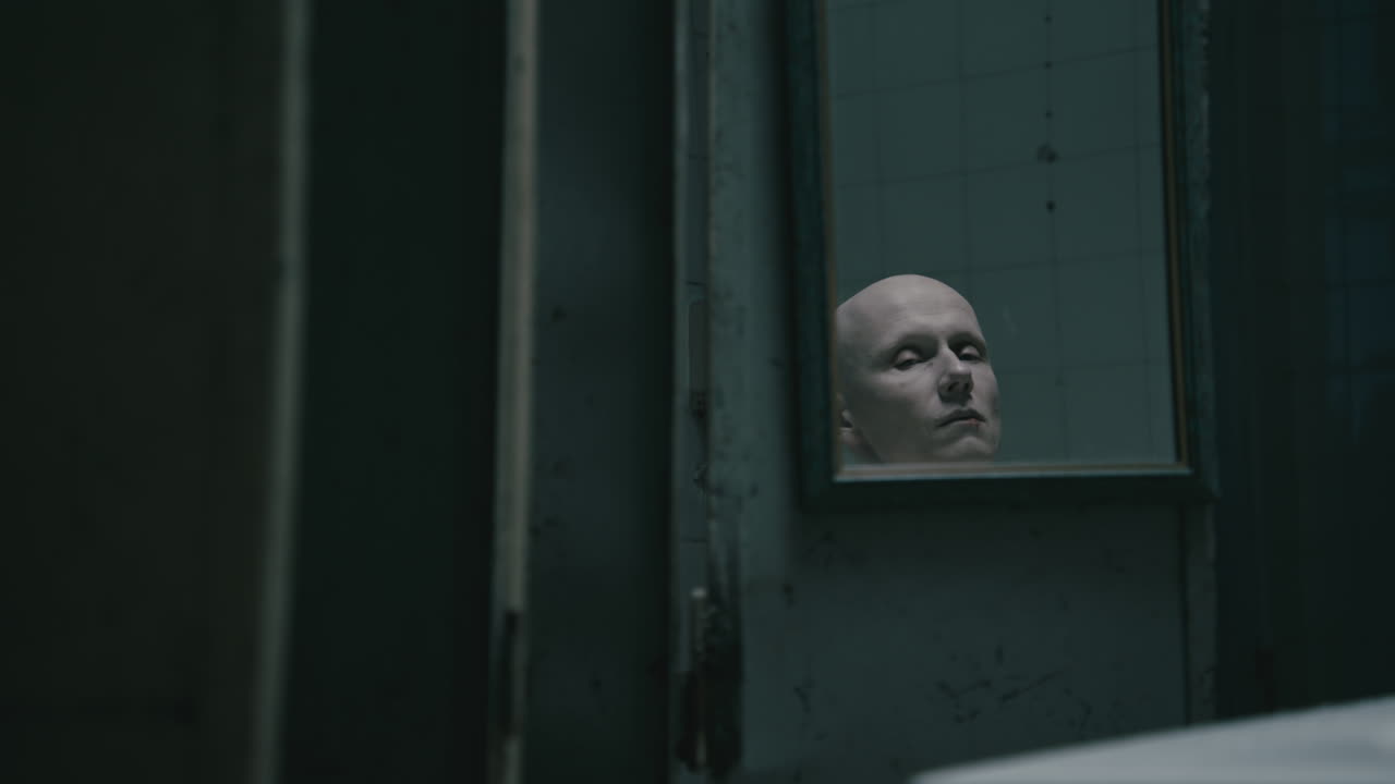 Pale bald skinny man looks into the mirror in the creepy old bathroom at night