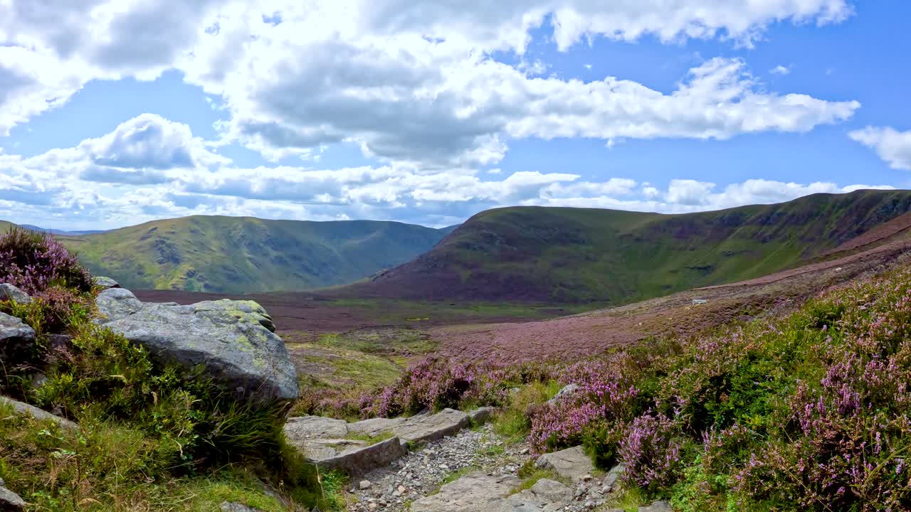 Camera moves steadily along a rocky footpath bordered by heather, revealing expansive Glen Clova hills under bright daylight and partly cloudy skies