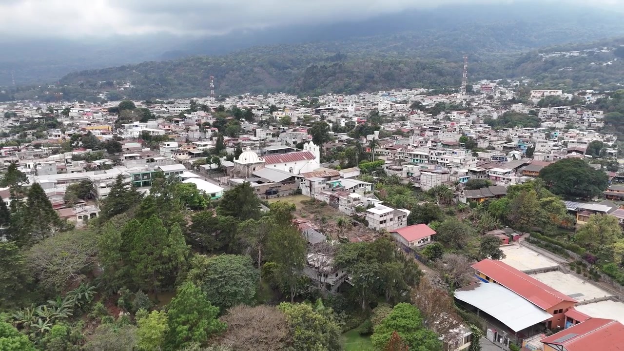 Vista aerea con drone su San Lucas, Guatemala, rivela quartieri tropicali, palme, il vulcano circostante e montagne boscose mentre il volo si sposta nel vivace paesaggio urbano.