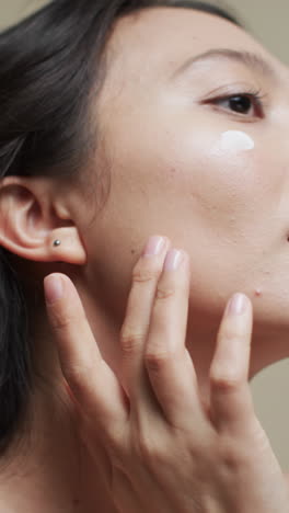 mujer asiática con cabello oscuro aplicando crema en su cara sobre un fondo beige, cámara lenta