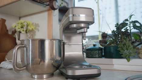 close up of silver electric stand blender with stainless mixing bowl on kitchen counter beside immersion blender and decorative flower vases with potted plants near window