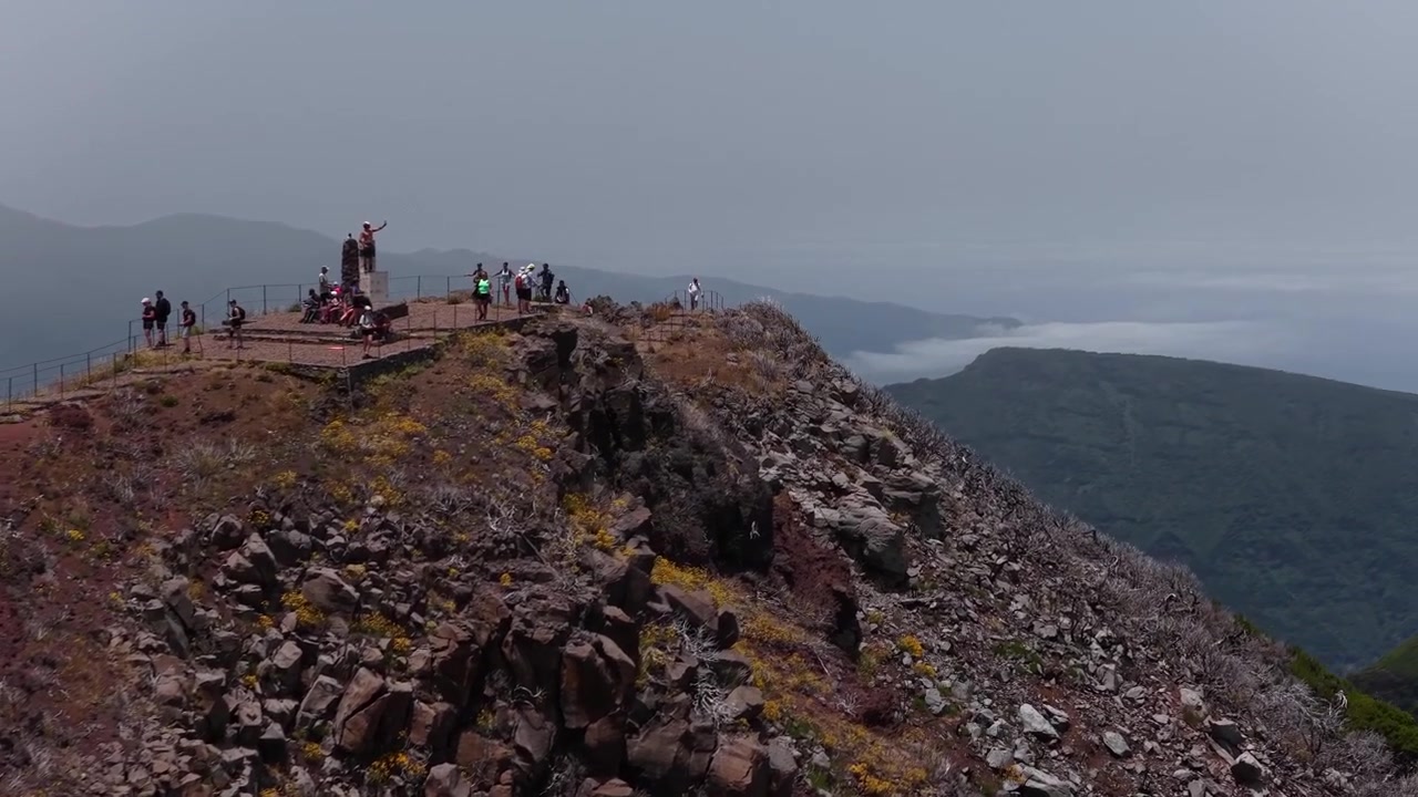 Drone footage of hikers at Pico do Areeiro summit viewpoint on Madeira’s iconic PR1 trail, revealing rugged cliffs, rocky textures, dramatic skies, and sweeping panoramic mountain vistas.