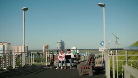 two students seated outdoors on benches, boy typing on laptop while girl points at something in her book, with an urban backdrop of buildings, railing, and greenery under clear blue sky