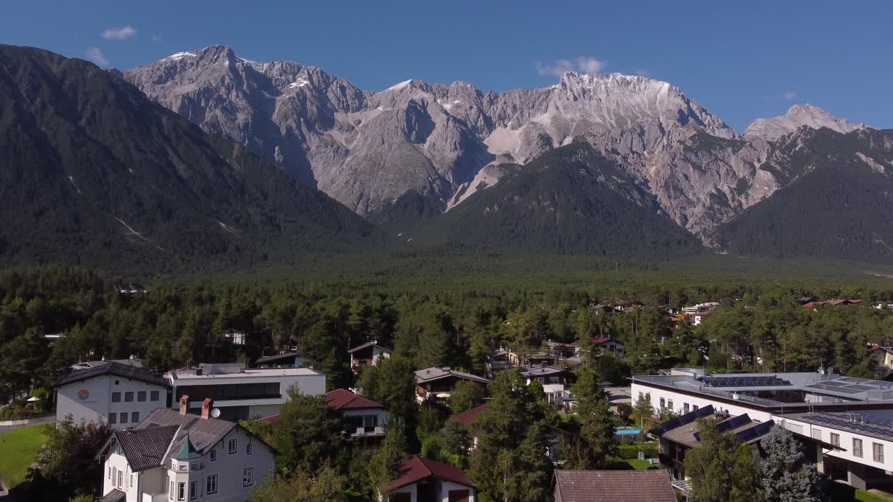 drone elevándose sobre un pequeño pueblo en los alpes con enormes montañas en la parte de atrás en un día soleado