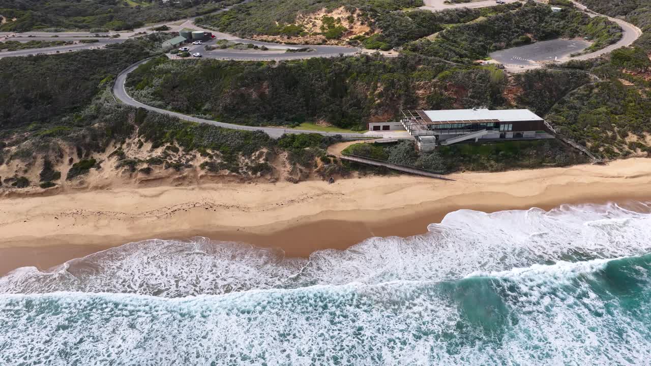 Drone glides above cliffside building, sandy beach, and crashing ocean waves in daylight