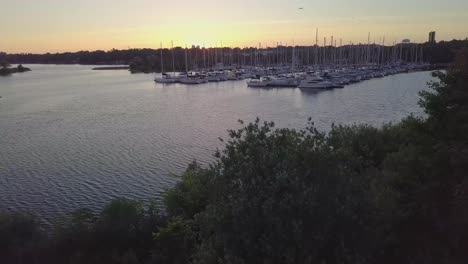 Toma-Aérea-Descendente-De-La-Puesta-De-Sol-Del-Puerto-Deportivo-Del-Club-Náutico-De-Velero-En-Los-Toboganes-De-La-Bahía-Del-Lago-A-La-Izquierda-Entre-árboles-Verdes-Con-El-Horizonte-De-Los-Edificios-De-La-Ciudad-En-El-Fondo-En-Toronto,-Ontario,-Canadá