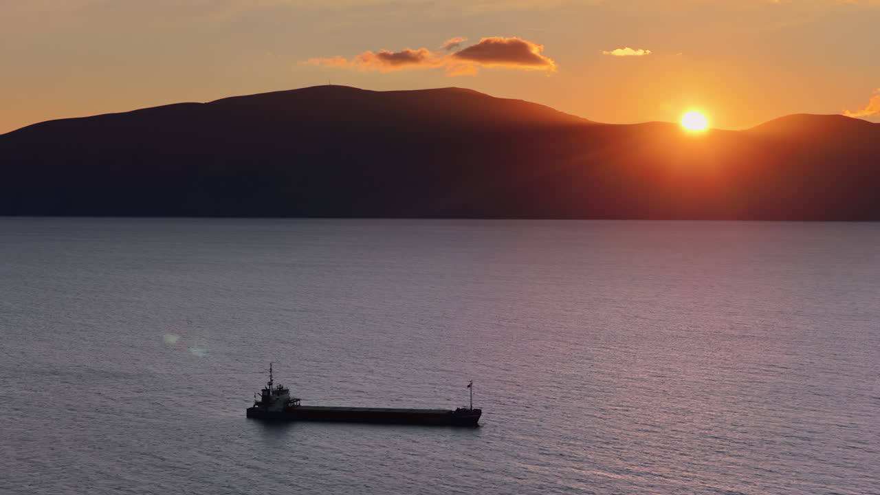 Aerial drone view of a lone fisherman rowing his small wooden boat during sunset in Vlora Bay, Albania, with soft orange skies, calm waters, and distant silhouettes of hills and clouds