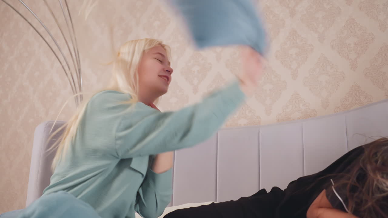 Close up of two college girls joyfully engaging in playful pillow fight on bed, one girl swings blue pillow with smile, other defends while laughing