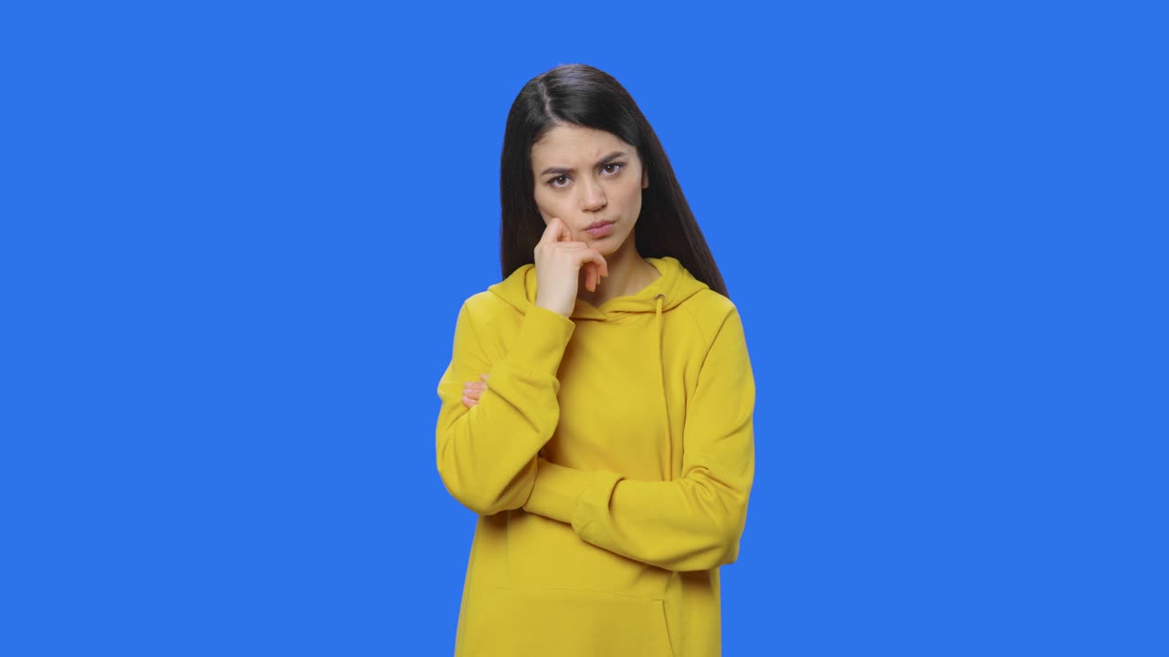 retrato de morena está pensando en algo, y luego una idea que viene a ella. mujer joven en sudadera amarilla posando en el estudio con fondo de pantalla azul. cerca. cámara lenta lista 59.94fps