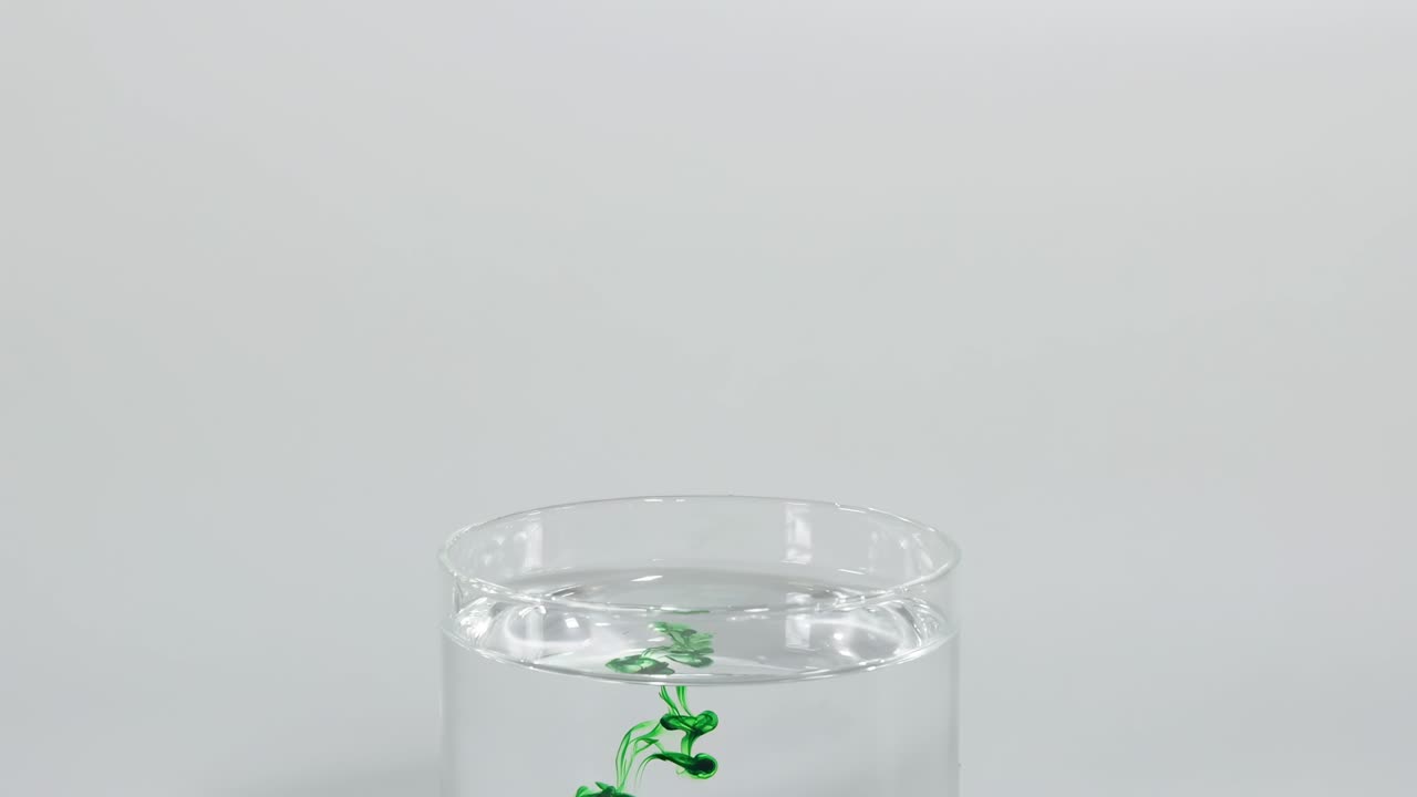 A dropper releases green dye into clear water, creating swirling patterns as it disperses.