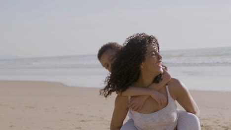 beautiful young american mother carrying her little son on her back while spending time at the beach on a sunny day