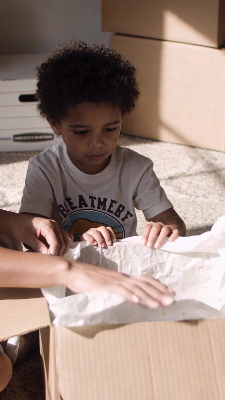 Video verticale: Adulto che si protende e guida un bambino con una maglietta bianca, sollevando un foglio per controllare gli articoli della scatola a casa