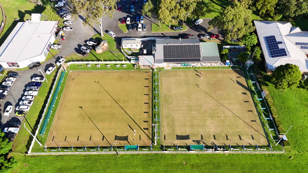 Drone footage captures two empty bowling fields surrounded by greenery and parked cars under bright sunlight