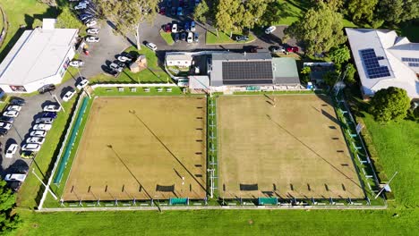 drone footage captures two empty bowling fields surrounded by greenery and parked cars under bright sunlight